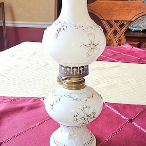 Op de afbeelding: Een witte antieke olielamp met een decoratief bloemmotief. De lamp heeft een ronde basis, een bolvormig middengedeelte en een hoge, uitlopende bovenkant. De metalen accenten zijn goud en brons. De lamp staat op een rood tafelkleed.