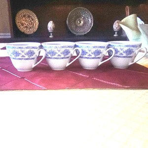Puede incluir: Cuatro tazas de té blancas con diseños florales azules y asas curvas dispuestas sobre un mantel rojo. En el fondo se ven platos y adornos decorativos, lo que sugiere un tema vintage o antiguo.