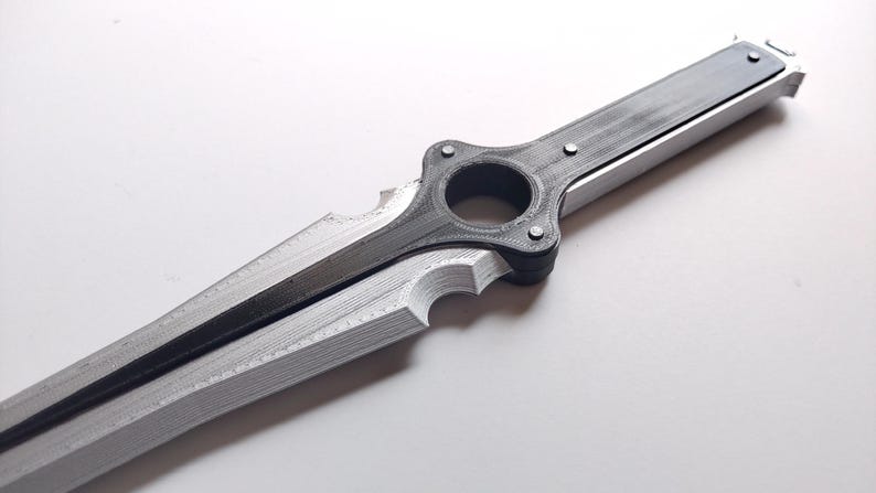 May include: A silver and black sword with a unique design. The blade is silver, and the handle and central ring are black. The sword has a distinctive shape with a circular opening near the handle and a notched edge.