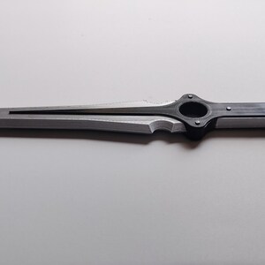 May include: A silver and black dagger with a sharp, pointed blade. The handle features a circular design and is connected to the blade with a decorative element. The dagger is lying on a white surface.