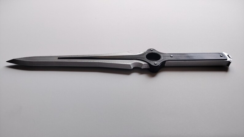 May include: A silver and black dagger with a sharp, pointed blade. The handle features a circular design and is connected to the blade with a decorative element. The dagger is lying on a white surface.