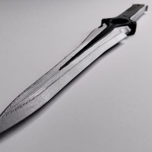 May include: A grayscale image of a decorative dagger. The blade is silver with a textured surface and a central groove. The handle is black and features a circular design. The dagger is lying on a white surface.