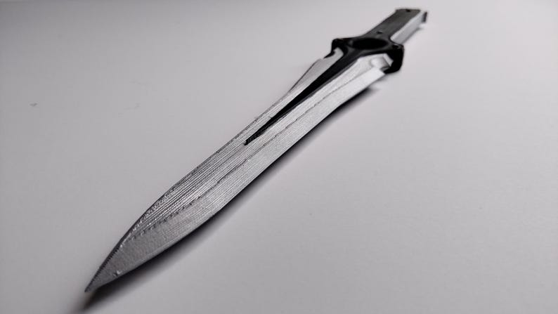 May include: A grayscale image of a decorative dagger. The blade is silver with a textured surface and a central groove. The handle is black and features a circular design. The dagger is lying on a white surface.