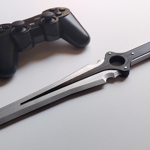May include: A black PlayStation 3 controller and a silver and black decorative sword are displayed on a white surface. The sword has a detailed handle and a sharp, pointed blade, suggesting a fantasy or gaming theme.
