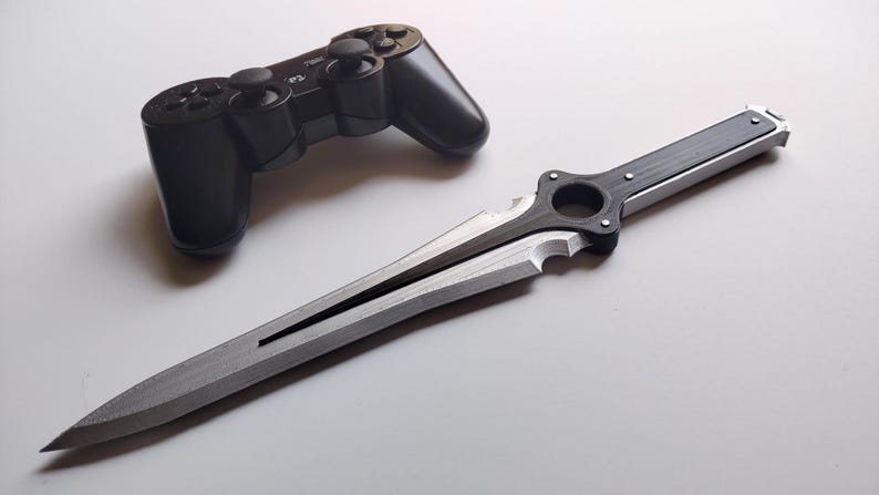 May include: A black PlayStation 3 controller and a silver and black decorative sword are displayed on a white surface. The sword has a detailed handle and a sharp, pointed blade, suggesting a fantasy or gaming theme.