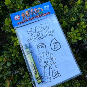 May include: A white coloring page with a Roblox character and the text "KALI TURNS 6". The page is in a clear plastic bag with two crayons, one green and one blue.