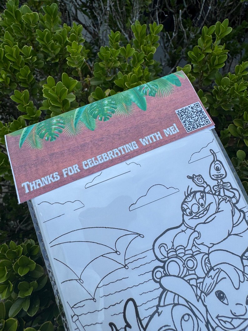 May include: A clear plastic coloring book with a brown and green decorative top. The top has the text "THANKS FOR CELEBRATING WITH ME!" and a QR code. The coloring book features a black and white cartoon illustration of characters.