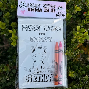 May include: A white coloring page with a black and white cow illustration and the text "Holy Cow It's Emma's Birthday". The page is packaged with two crayons, one pink and one orange.