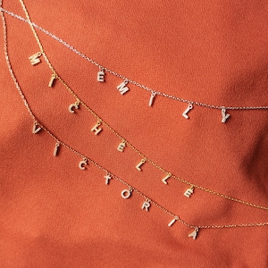 Könnte beinhalten: Fünf goldene Halsketten mit Buchstabenanhängern, die die Namen "VICTORIA", "HELLER", "EMILY" und "MICHAEL" buchstabieren. Die Halsketten liegen auf einem braunen Stoffhintergrund.