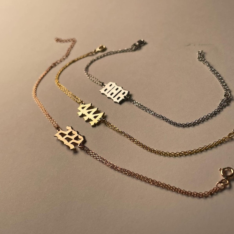 Puede incluir: Tres pulseras de cadena de oro con diferentes n&uacute;meros en los dijes. Los n&uacute;meros son 222, 444 y 888. Los dijes est&aacute;n en una fuente g&oacute;tica.