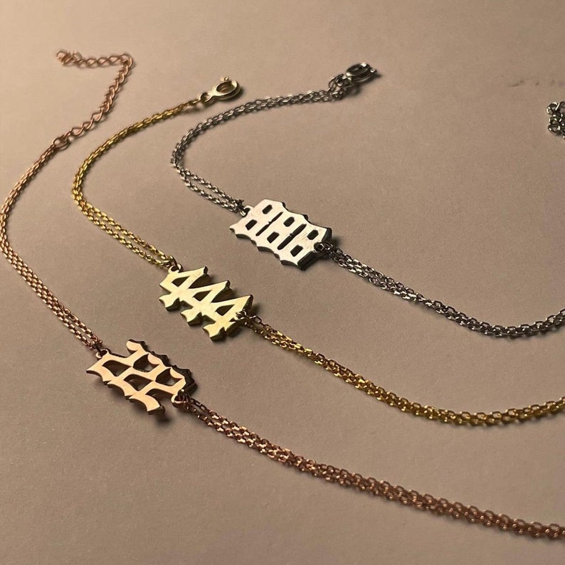 Puede incluir: Tres pulseras de cadena de oro con diferentes dijes de n&uacute;meros. Los dijes est&aacute;n en una fuente g&oacute;tica y dicen "222", "444" y "888".