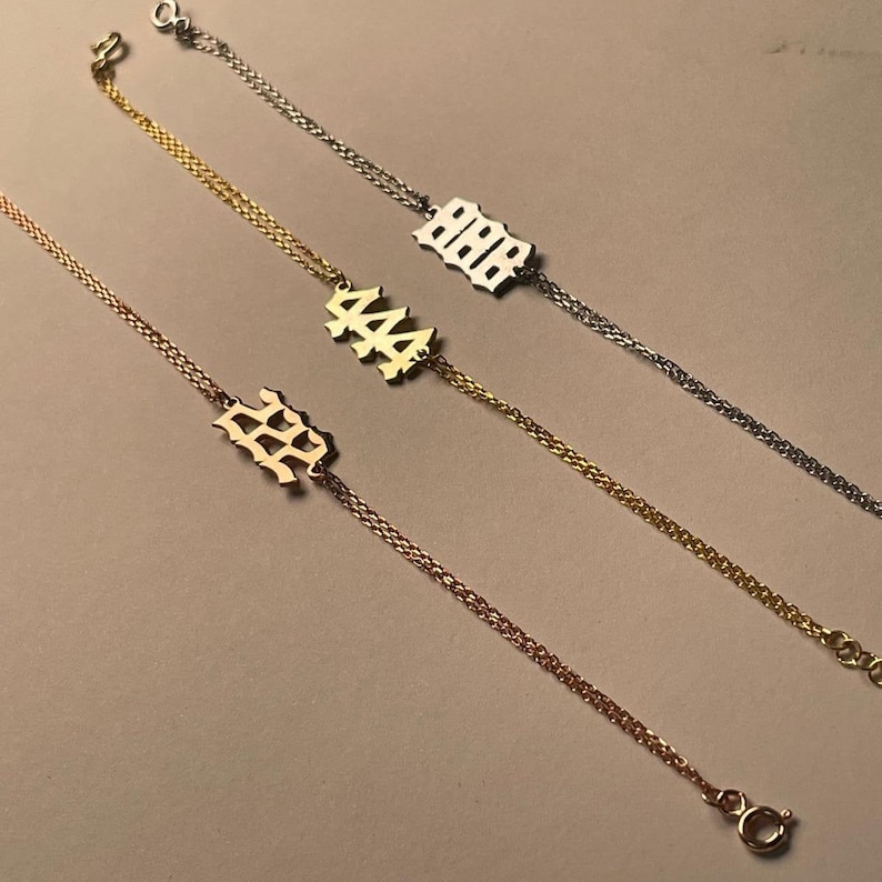 May include: Three gold chain bracelets with different numbers on the charms. The numbers are 222, 444, and 888. The charms are in a gothic font.