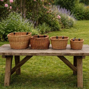 May include: Four brown wicker baskets of different sizes are displayed on a wooden table in a garden. The baskets have black leather straps. The table is made of weathered wood. The background features green foliage and pink and purple flowers.