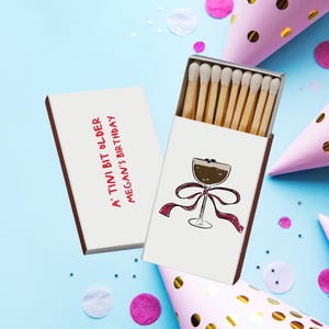 May include: A matchbox with a cocktail illustration and the text "A tini bit older Megan's Birthday" is open, revealing wooden matches. The box is next to a closed matchbox with the same text. Party hats and confetti are scattered on a blue background.