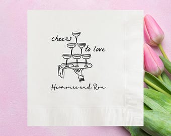May include: White square napkin with a black illustration of champagne glasses stacked on a tray, held by a hand. The words "cheers to love" and the names "Hermonie and Ron" are written on the napkin. Two pink tulips are on the right side.