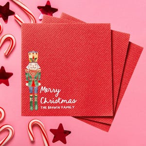 May include: Red Christmas-themed napkins with a watercolor nutcracker illustration and the text "Merry Christmas". The napkins are surrounded by candy canes and star-shaped decorations on a pink background.