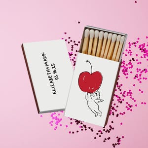 May include: A white matchbox with a red cherry illustration and the text "ELIZABETH+MARK 03.16.25" on a pink background. The matchbox is open, revealing wooden matches. Pink heart-shaped confetti is scattered around.