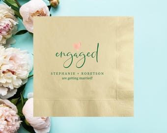 May include: A beige paper napkin with the word "engaged" in green script, a pink heart, and the names "Stephanie + Robetson" with the text "are getting married!" in green. The napkin is surrounded by white and pink peonies on a light blue background.