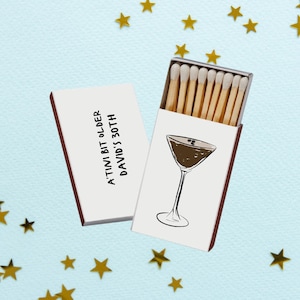 May include: Two white matchboxes with red edges, one open and one closed, against a light blue background with gold star confetti. The open box displays matches and a cocktail glass illustration. Text on the closed box reads: "A'TINI BIT OLDER DAVID'S 30TH."