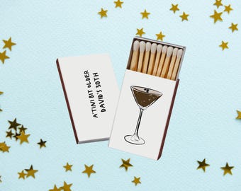 May include: Two white matchboxes with red edges, one open and one closed, against a light blue background with gold star confetti. The open box displays matches and a cocktail glass illustration. Text on the closed box reads: "A'TINI BIT OLDER DAVID'S 30TH."