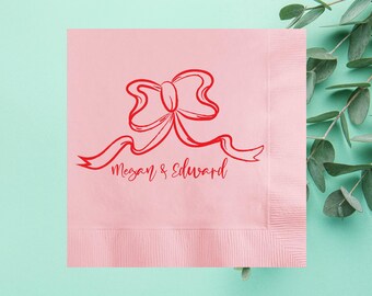May include: A pink paper napkin with a red bow design and the names "Megan & Edward" in script. The napkin has a textured surface and a decorative border. The background is a light teal color.