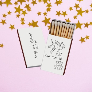 May include: Matchbox with wooden matches, open to reveal the match heads. The box is white with a red border and features a drawing of an angel and the words "cin cin". The box also has the names "Emily and Richard" and the date "10.18.24". Gold star confetti on a pink background.