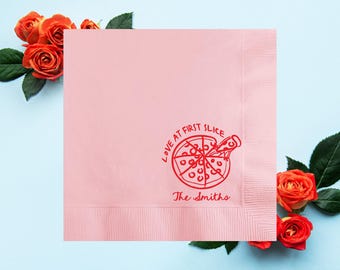 May include: A pink napkin with a red pizza graphic and the text "Love at First Slice" and "The Smiths." The napkin is surrounded by orange roses and green leaves on a light blue background.