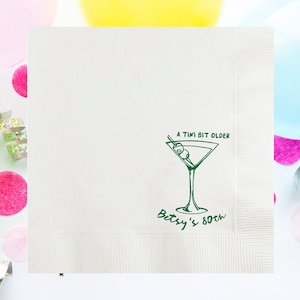May include: White cocktail napkin with a green martini glass illustration and the text "A tini bit older" and "Betsy's 80th". The napkin is surrounded by colorful balloons and confetti.