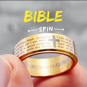 May include: A gold-colored spinning ring with a black and white image of a cross and bible verses. The ring has the words "BIBLE" and "SPIN" printed on it. The text on the ring reads: "Against thee shall prosper. And of a good courage. For I am with you, saith the Lord. I can do all things through Christ which strengtheneth me: Trust in the Lord with all thine heart; and lean not unto thine own understanding. In all thy ways acknowledge him, and he shall direct thy paths.  Man of God.  SH.1:9/1TIM 6:11-12/JER.29:11"