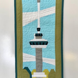 Op de afbeelding: Een groene en witte ingelijste gequilte wandhanger met een afbeelding van een hoge toren met een grijze basis en een groen-witte top. De toren staat tegen een blauwe lucht met witte wolken.