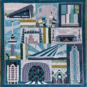 Op de afbeelding: Een quilt met een patchwork-ontwerp van verschillende gebouwen en stadsgezichten in tinten blauw, grijs en groen. De quilt is omlijst met een turquoise rand.