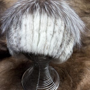 Op de afbeelding: Een luxe wintermuts met een bontrand. De muts heeft een mix van witte, grijze en bruine vacht, waardoor een textuur ontstaat. Het wordt gepresenteerd op een draadstandaard tegen een bruine vachtachtergrond.
