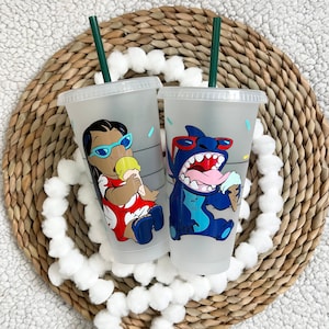 May include: Two clear plastic reusable cups with lids and straws. The cups feature a cartoon character design. One cup has a blue character with a red nose and sunglasses eating ice cream. The other cup has a brown character with blue eyes and sunglasses eating ice cream.