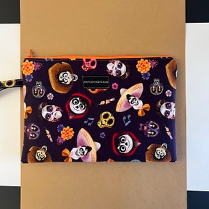 May include: A black zippered pouch with a colorful print of characters from the movie Coco. The print features skeletons with flowers, musical notes, and the text "100% HOMEMADE".