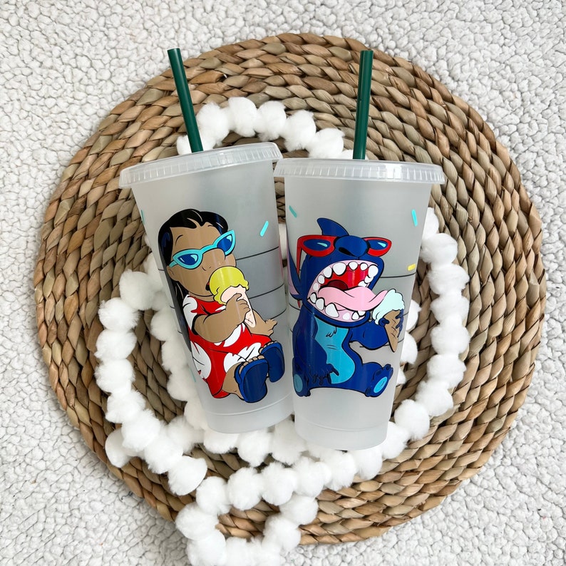 May include: Two clear plastic reusable cups with lids and straws. The cups feature a cartoon character design. One cup has a blue character with a red nose and sunglasses eating ice cream. The other cup has a brown character with blue eyes and sunglasses eating ice cream.
