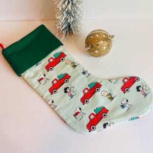 May include: A Christmas stocking with a green cuff and a light green body. The body is patterned with red trucks carrying Christmas trees, Snoopy, Charlie Brown, and other Peanuts characters. A small Christmas tree and a gold glitter ornament are in the background.