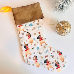 May include: A Christmas stocking with a brown cuff and a white body patterned with cartoon characters in red, tan, and blue. A gold ribbon is attached to the cuff. A gold glitter ornament and a small decorative tree are in the background.