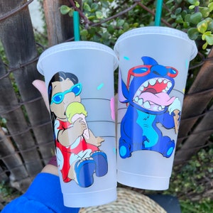 May include: Two white reusable cups with lids and straws. One cup features a cartoon image of Lilo from Lilo & Stitch wearing a red dress and sunglasses, holding an ice cream cone. The other cup features a cartoon image of Stitch wearing sunglasses and holding an ice cream cone.