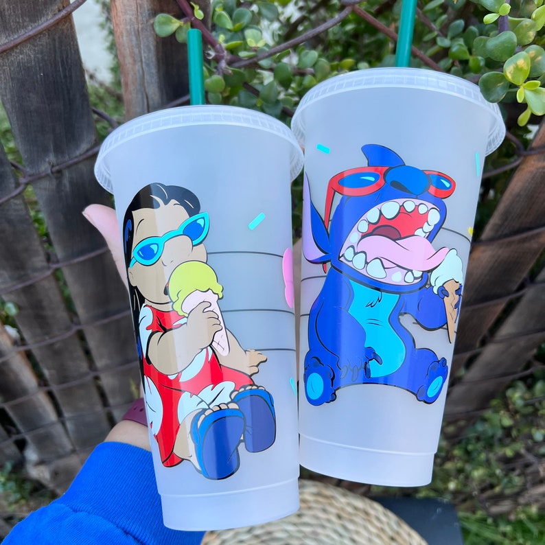 May include: Two white reusable cups with lids and straws. One cup features a cartoon image of Lilo from Lilo & Stitch wearing a red dress and sunglasses, holding an ice cream cone. The other cup features a cartoon image of Stitch wearing sunglasses and holding an ice cream cone.