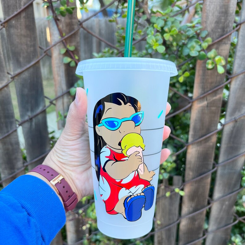 May include: A white reusable cup with a design of a cartoon character with black hair, blue sunglasses, and a red dress eating an ice cream cone. The character is Lilo from the Disney movie Lilo & Stitch.