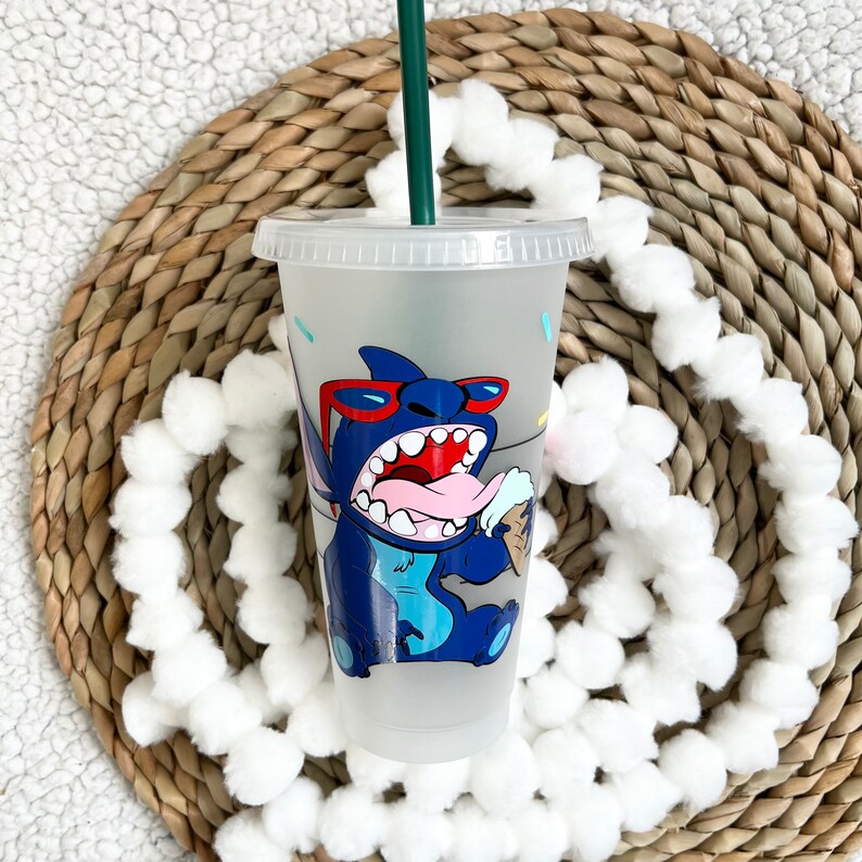 May include: A clear plastic reusable cup with a blue and white cartoon character design. The character is wearing red sunglasses and holding an ice cream cone. The cup has a green straw.