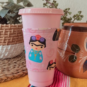 May include: Pink reusable cup with a pink and white fabric sleeve featuring a cartoon image of Frida Kahlo. The sleeve has a heart pattern and a white border.