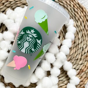 May include: A white reusable cup with a green Starbucks logo and colorful ice cream cone designs.