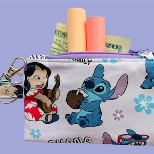 May include: A white zippered coin purse featuring cartoon characters Lilo and Stitch. The purse has a purple zipper and a matching wrist strap. Inside are two lipsticks and some cash. The background is a light purple.