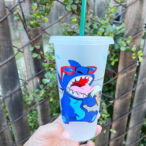 May include: A frosted blue reusable cup with a cartoon image of Stitch from Lilo & Stitch wearing sunglasses and eating an ice cream cone. The cup has a green straw.