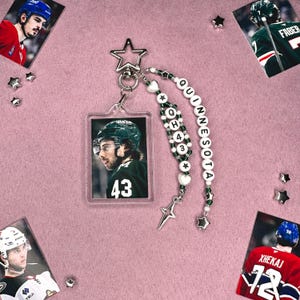 May include: A clear acrylic keychain featuring a hockey player in a green jersey with the number 43. The keychain is adorned with a beaded strap spelling "MINNESOTA" and decorative charms. Additional hockey player photos and star-shaped accents are scattered around.