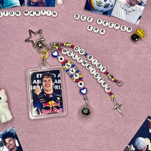 May include: A clear acrylic keychain featuring a photo of a smiling person in a racing suit. The keychain has beaded accents with the words "DU BIST WELTMEISTER" and "V5". Additional charms include a star and a disco ball.