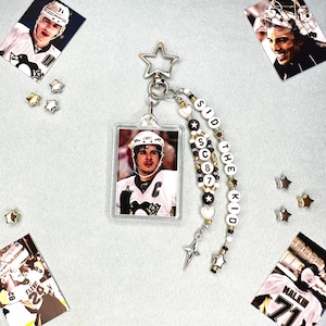 May include: A rectangular acrylic keyring featuring a photograph of a hockey player in a white jersey. The keyring includes a star-shaped clasp and two beaded strands with white letter beads spelling SID THE KID and SC 87, accented by small stars.
