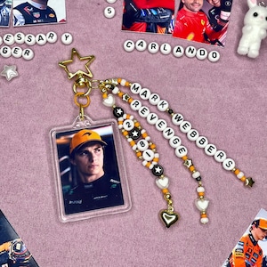 May include: A clear acrylic keychain featuring a photo of a race car driver, with a gold star charm and beaded accents. The beaded strands spell out "MARK WEBBERS REVENGE 28". The keychain includes gold heart and star charms.