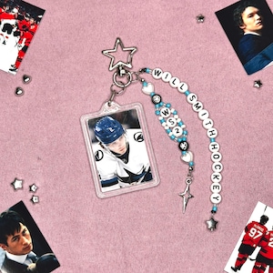 Könnte beinhalten: Ein klarer Acryl-Schlüsselanhänger mit einem Eishockeyspieler in blau-weißer Uniform. Der Schlüsselanhänger ist mit einem Perlenanhänger verziert, der "WILL SMITH HOCKEY" buchstabiert, und enthält stern- und herzförmige Charms. Der Hintergrund ist rosa.